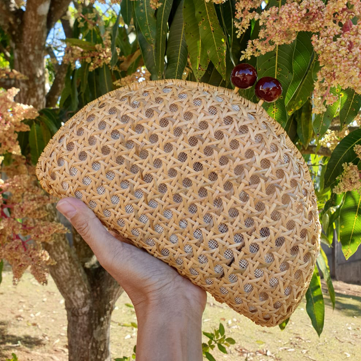 Mão segurando uma clutch de vime tecida com dois fechos vermelhos, cercada por folhas verdes e flores rosa.